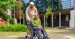 One man supports another man who is using a wheelchair. They are outside in a park-like setting on a summer day.