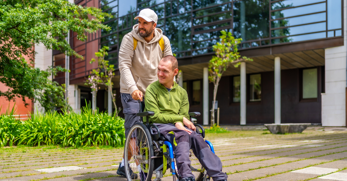 One man supports another man who is using a wheelchair. They are outside in a park-like setting on a summer day.