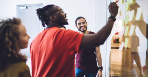 A black man wearing a red shirt points his pen at a whiteboard with sticky notes. Two other colleagues observe.