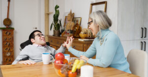 Man with an intellectual disability is in a wheelchair at a kitchen table, high-fiving his female support staff and smiling.