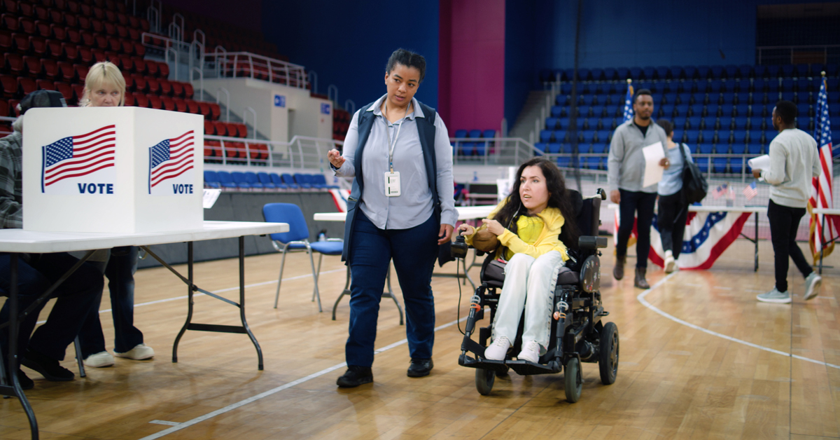 Woman using an electric wheelchair is in a gymnasium on election day, accompanied by a support staff member who is assisting her with voting.