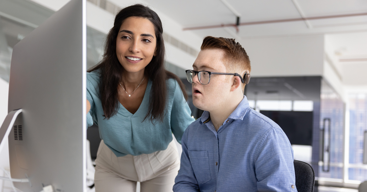 A female DSP supports a man with Down syndrome to use a computer. They are both at a desk in a modern office setting